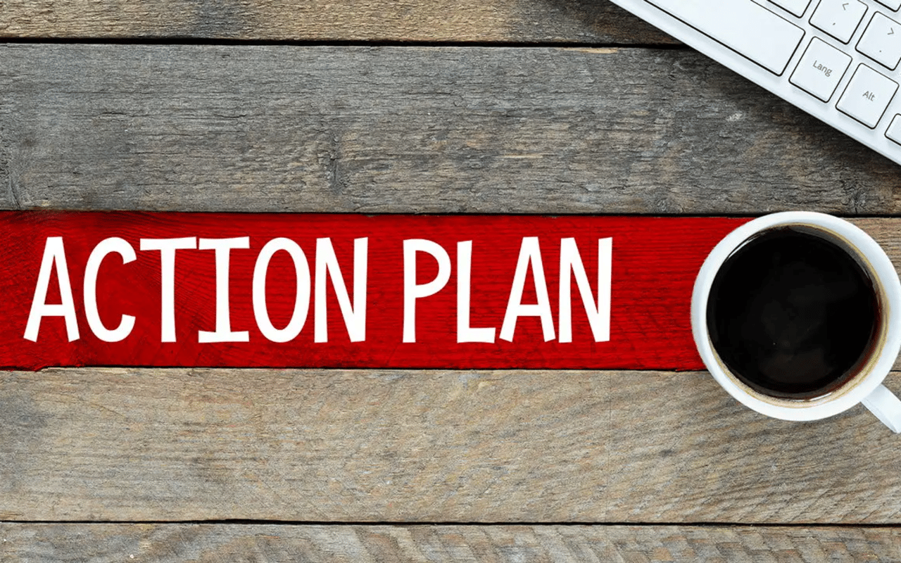 Action plan lettering on a red strip across a wooden desk, beside a keyboard and black coffee.