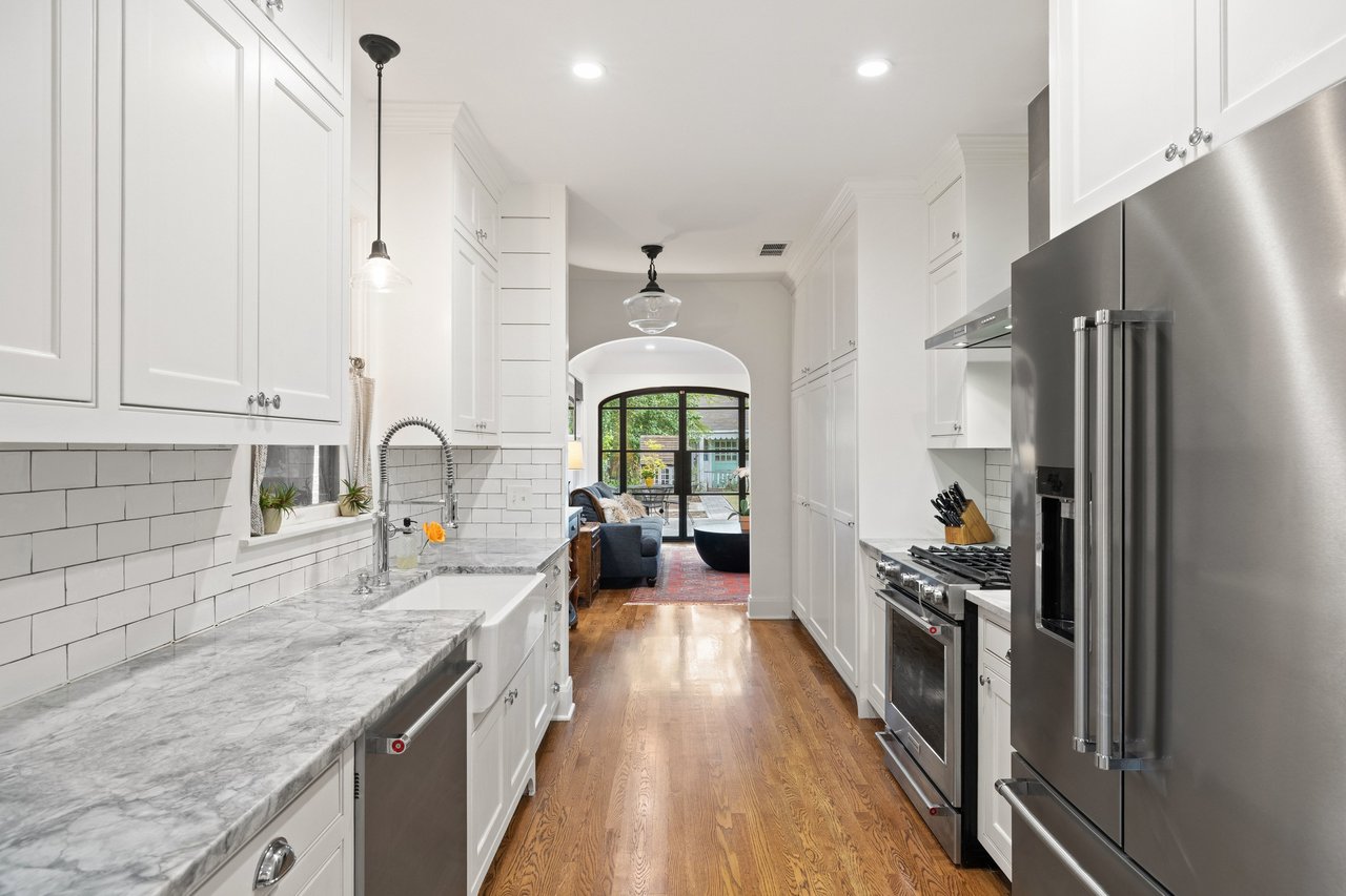 Kitchen with marble counters and farmhouse sink, looking through an archway to a furnished living area.