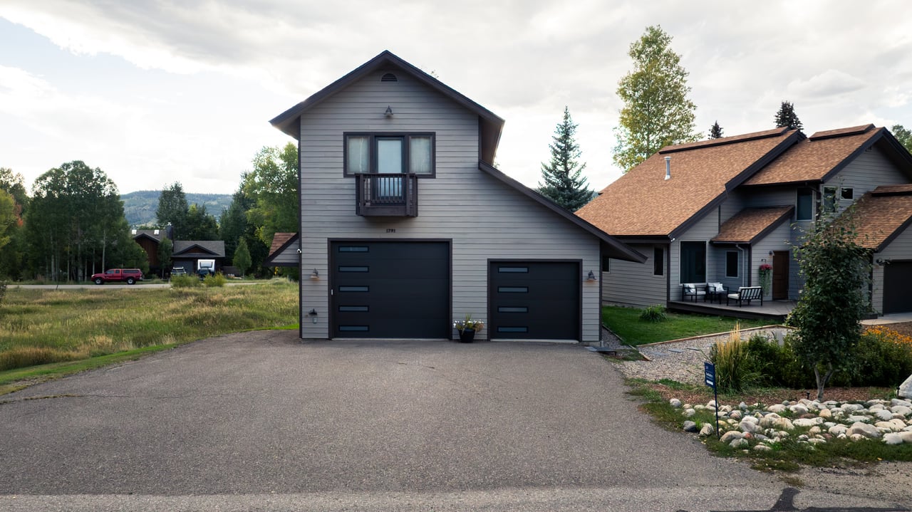Gray-sided property with two dark garage doors, a broad driveway, and an adjoining brown-roofed residence.