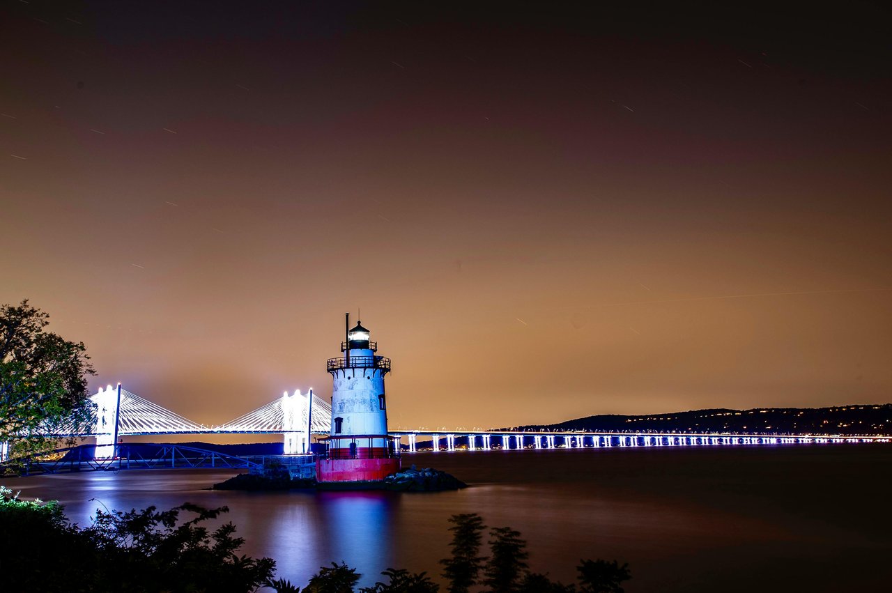 Lighthouse and illuminated bridge over the Hudson River at night.