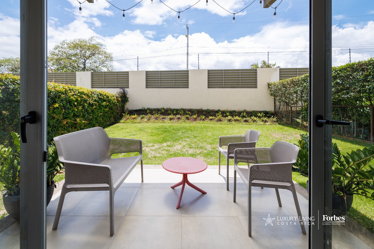 Furnished patio with three woven chairs and a red table overlooking a private lawn and garden.