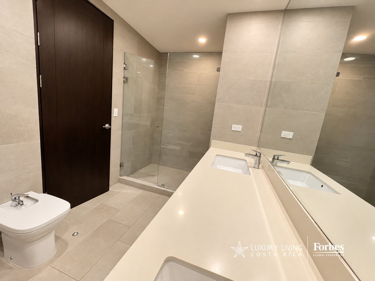 Modern tiled bathroom with a glass shower, toilet, and long cream vanity.