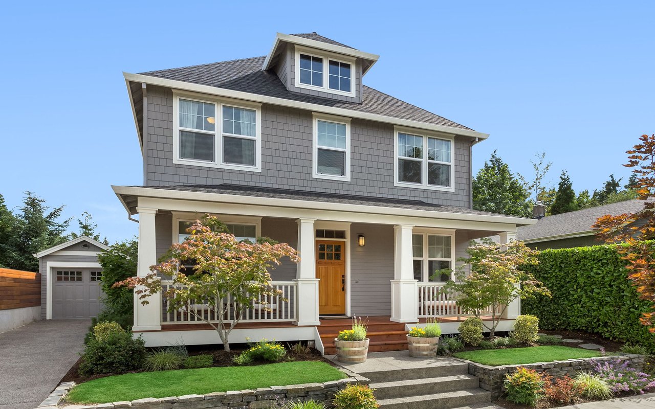 Gray two-story house with a covered porch, dormer window, side garage, and landscaped front yard.