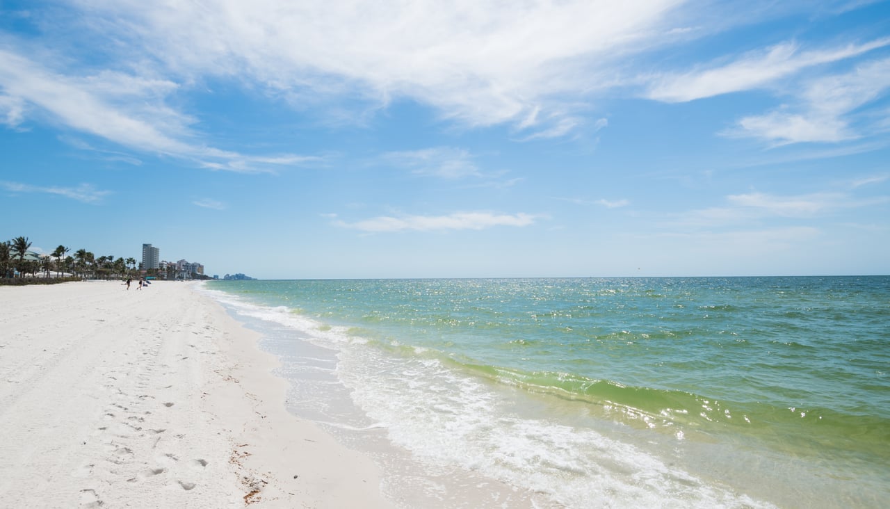 White-sand beach and blue-green water with distant beachfront buildings.