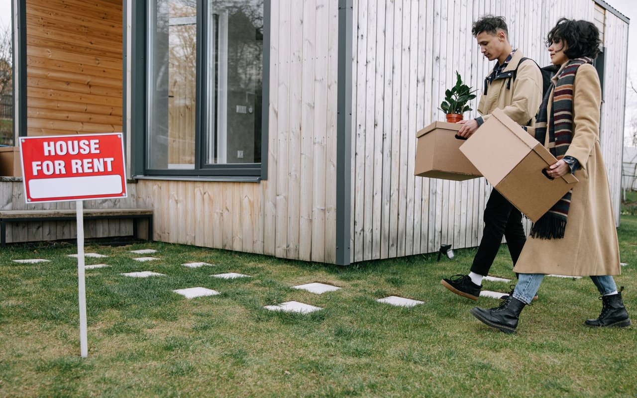 A couple carries moving boxes past a house with a “HOUSE FOR RENT” sign.