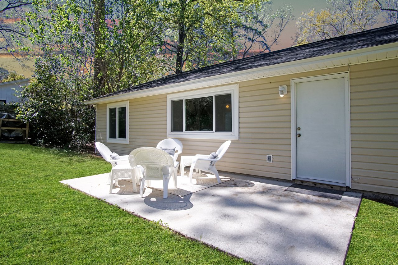 Small beige house with white trim, featuring a patio with white chairs and a table. Surrounded by green grass and trees, evokes a peaceful, sunny day.