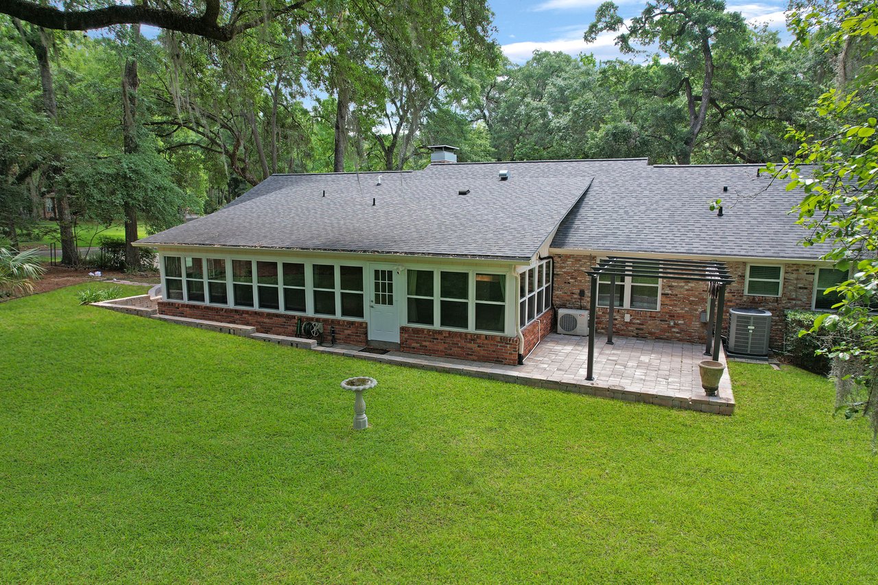 A single-story brick house with a large sunroom and a patio sits among lush green grass and mature trees, evoking a serene, natural setting.