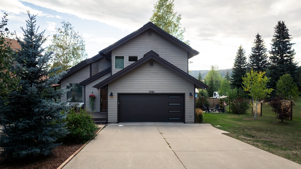 Gray multi-level house with a wide dark garage, concrete driveway, and mature evergreens.