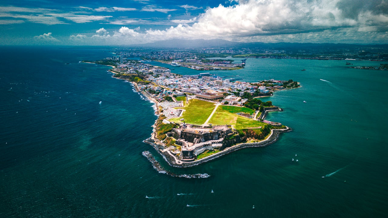 Aerial view of Old San Juan with El Morro fort, lush green open spaces, and vibrant city buildings surrounded by the blue Atlantic Ocean.