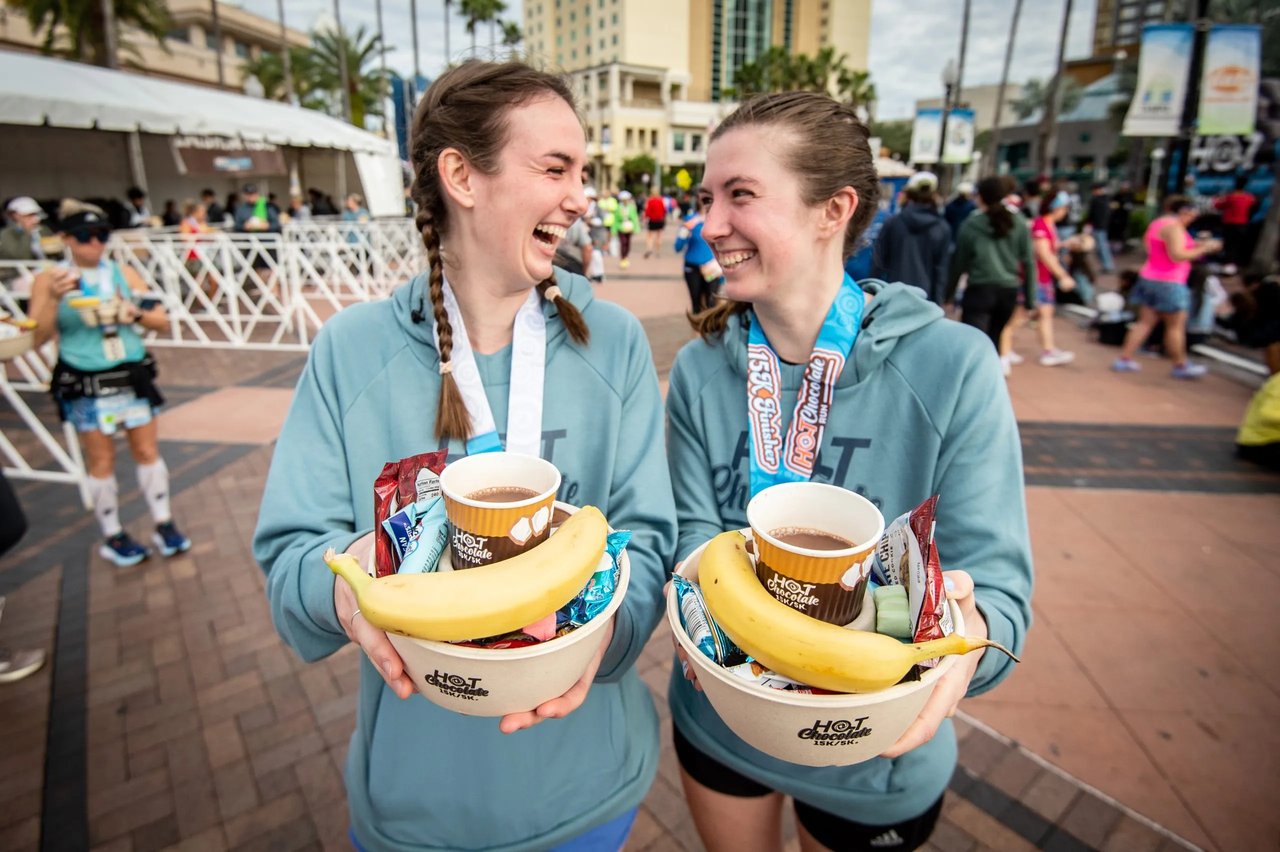 1. Optimized Alt-Description Two smiling runners holding "Hot Chocolate 15k/5k" finisher mugs with bananas and chocolate fondue after the race.