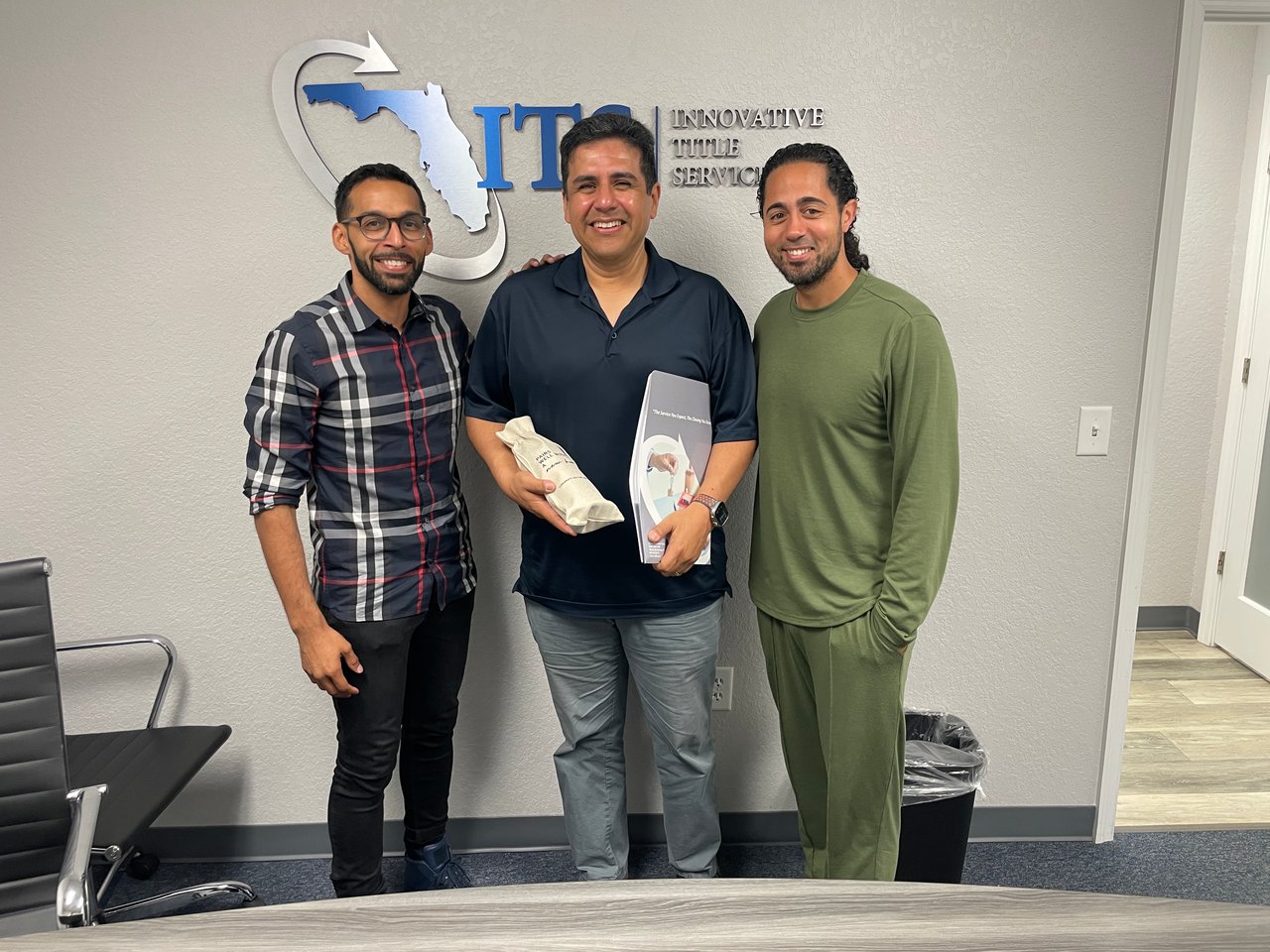 Three men pose in an office; the center man holds a wrapped gift and booklet.