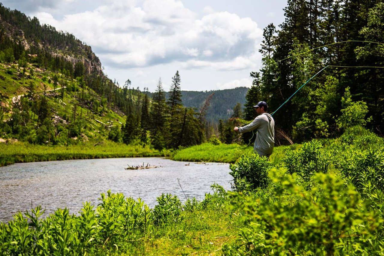 A fisherman casting a fly-fishing line into a pristine, clear mountain river surrounded by lush green hills.