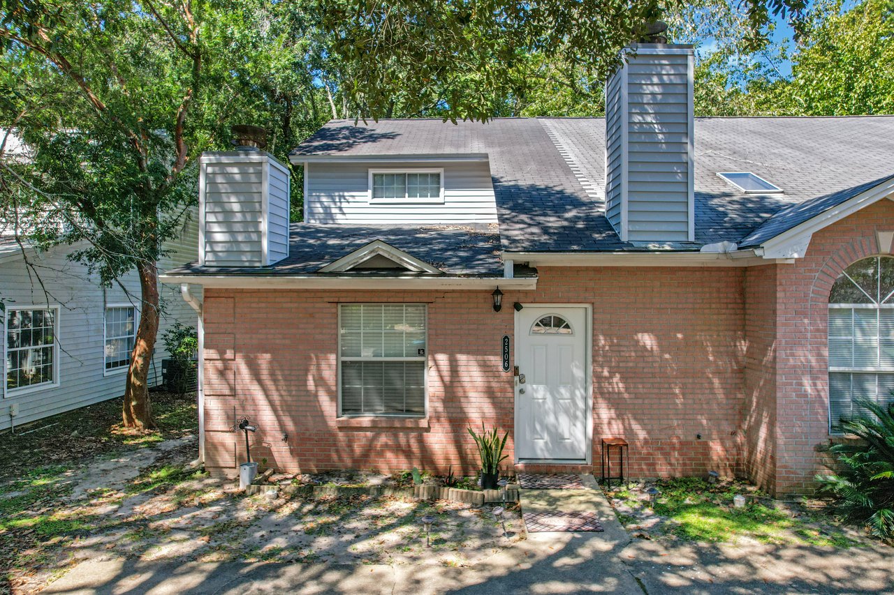 Straight-on view of the front exterior of a brick townhome at 2506 Nugget Lane in Tallahassee, Florida. The home features a white entry door, rectangular front window with blinds, and an arched window on the right. Two chimneys with gray siding rise above the roofline, with mature trees providing shade over the entry walkway and small landscaped area.