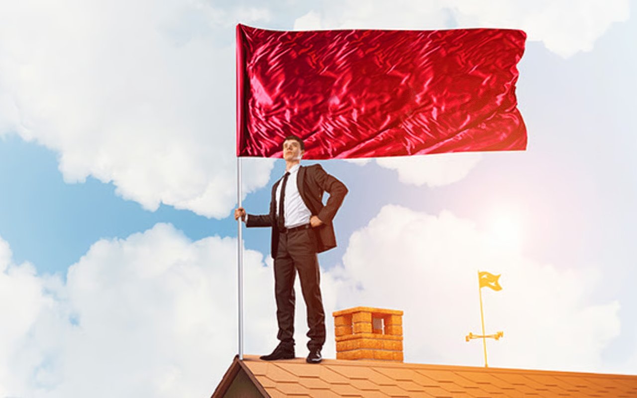 A suited man stands on a roof holding a large red flag against a cloudy sky.