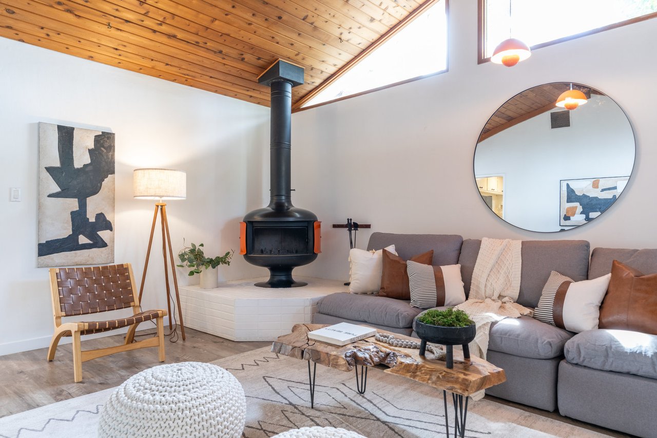 Mid-century modern living room with wood ceiling, gray sectional, round mirror, and black freestanding fireplace.