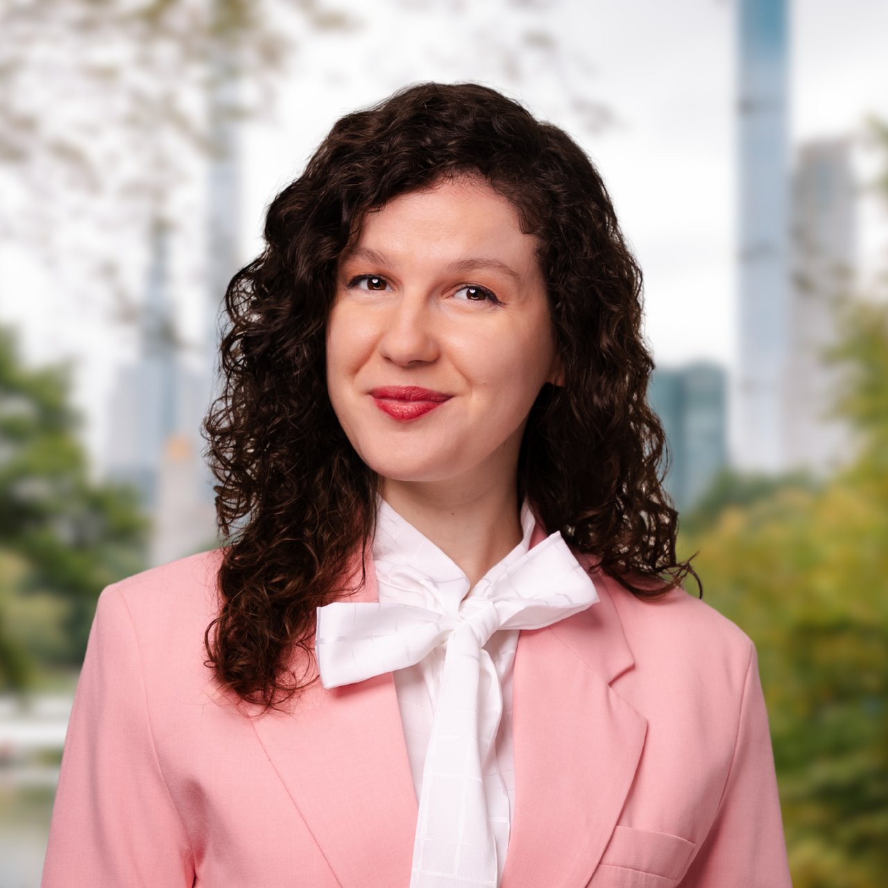 Tiziana Marku in a pink blazer and white bow blouse, against a softly blurred city backdrop.