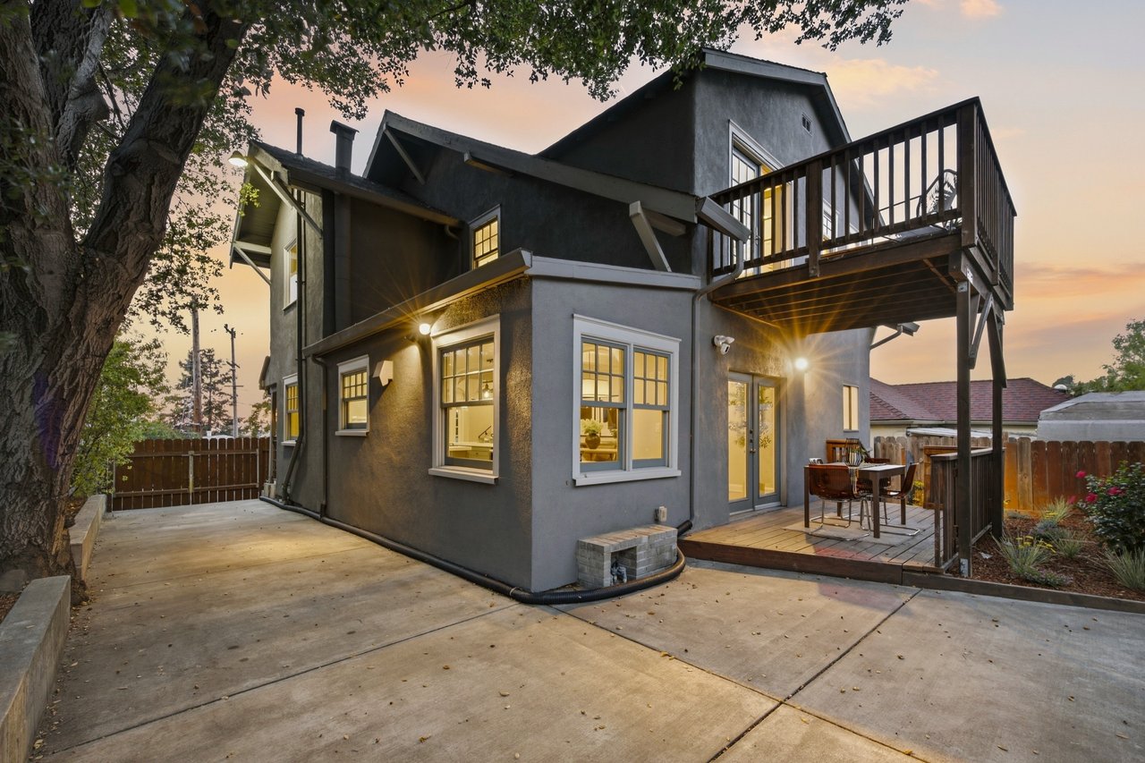 Two-story gray house at dusk with a raised wooden balcony, patio, and broad concrete driveway.