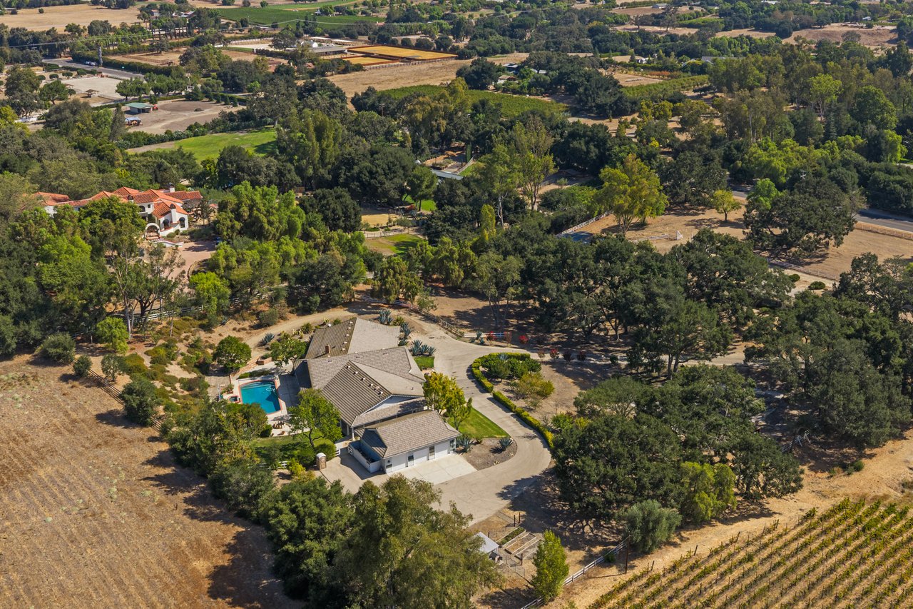 Aerial view of a rural home with a pool, mature trees, and vineyard rows.