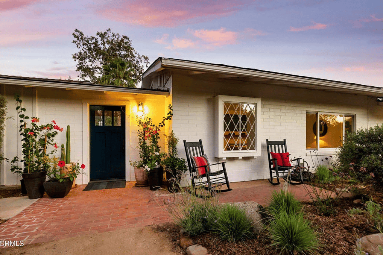 White brick home with a dark front door, brick patio, rocking chairs, and potted flowers at dusk.