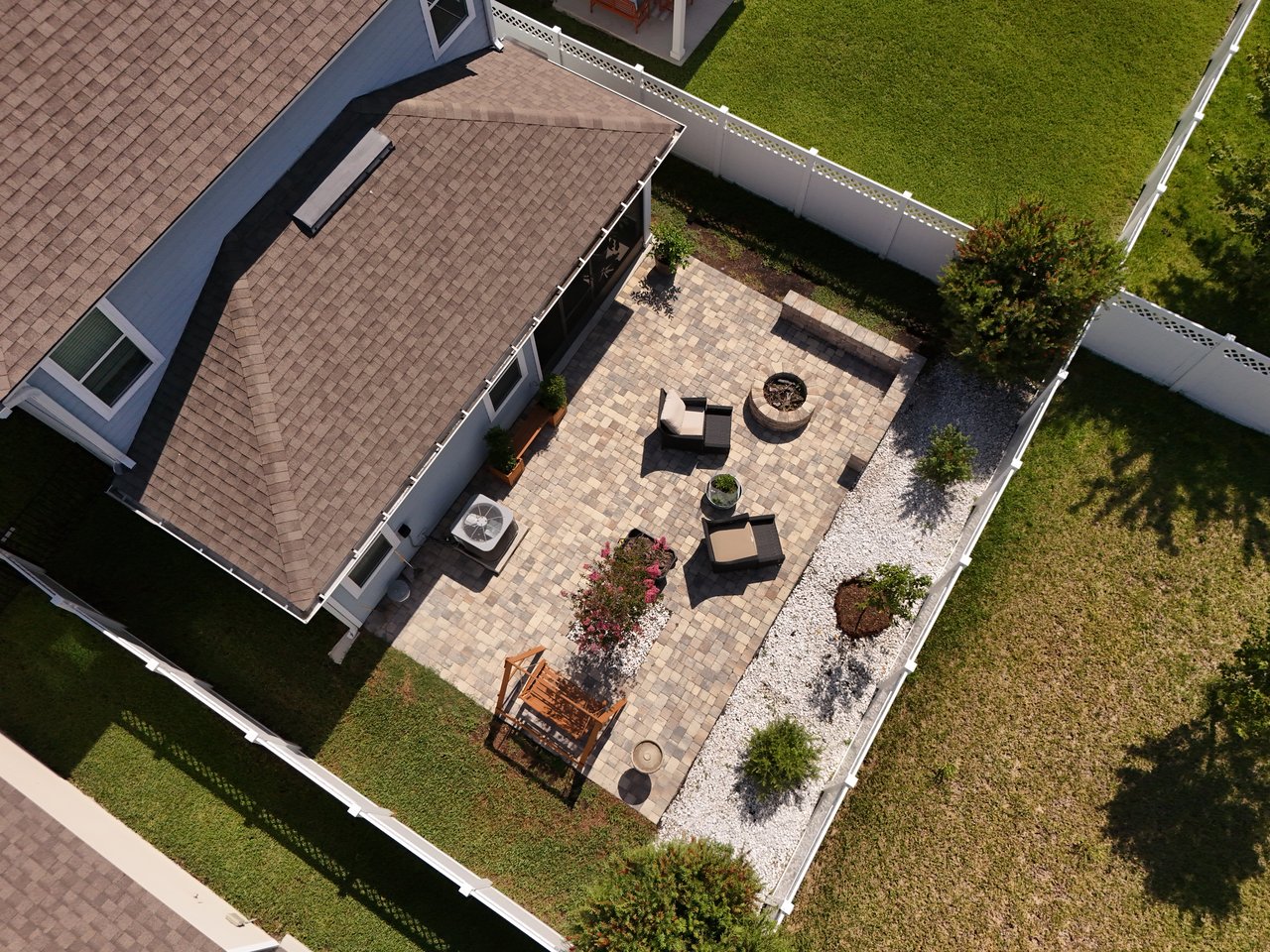 Aerial view of a paver backyard patio with seating, fire pit, and white fenced yard.