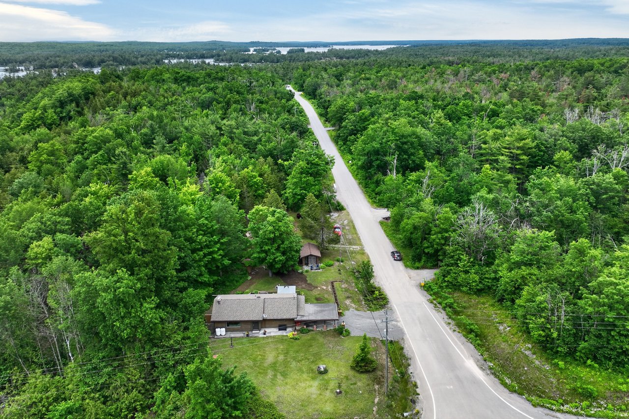 Aerial view of a house beside a rural road, surrounded by dense forest with distant water on the horizon.