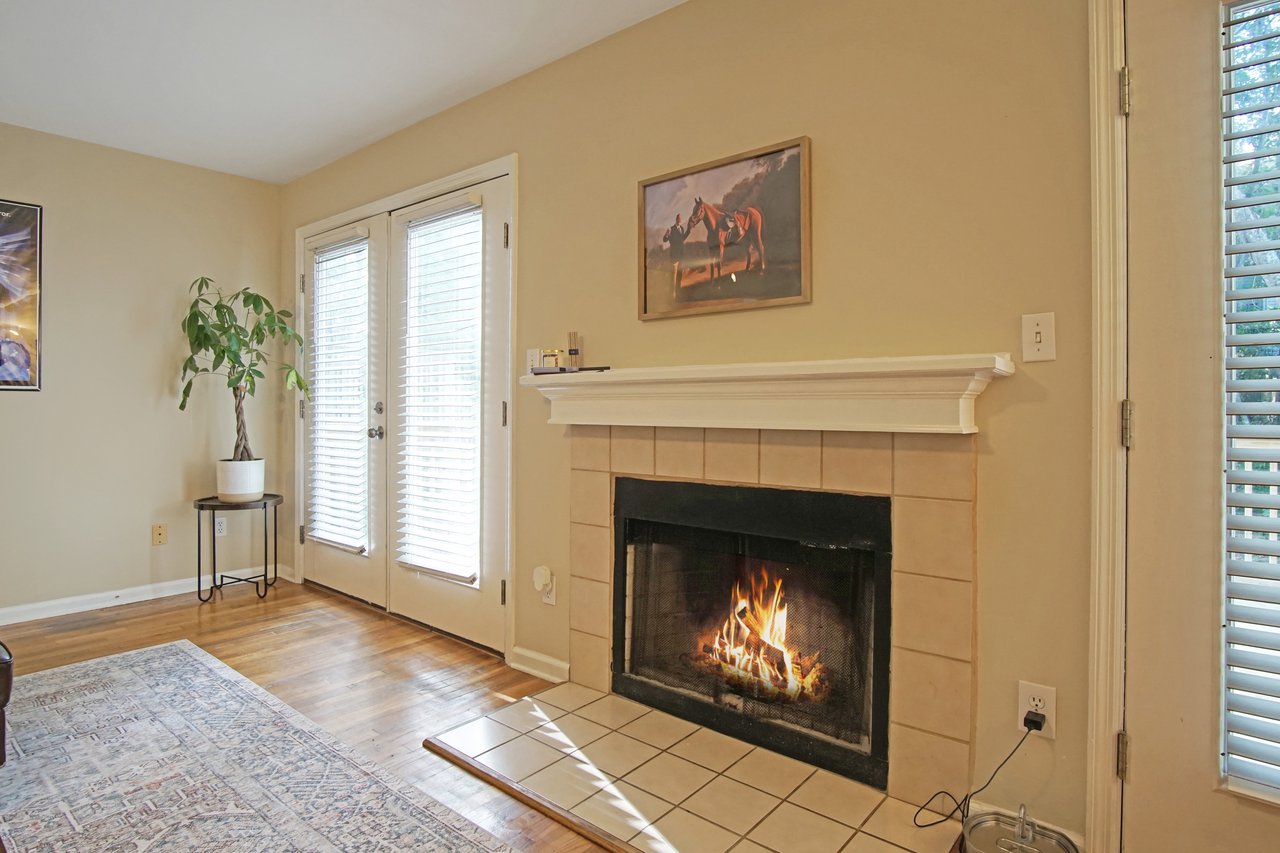 Living room fireplace at 529 Collinsford Road, Tallahassee, Florida, featuring a tile surround and white mantel. The area includes French doors with blinds that open to the back deck, a potted plant by the window, and a wall-mounted painting above the mantel, creating a cozy and inviting atmosphere.