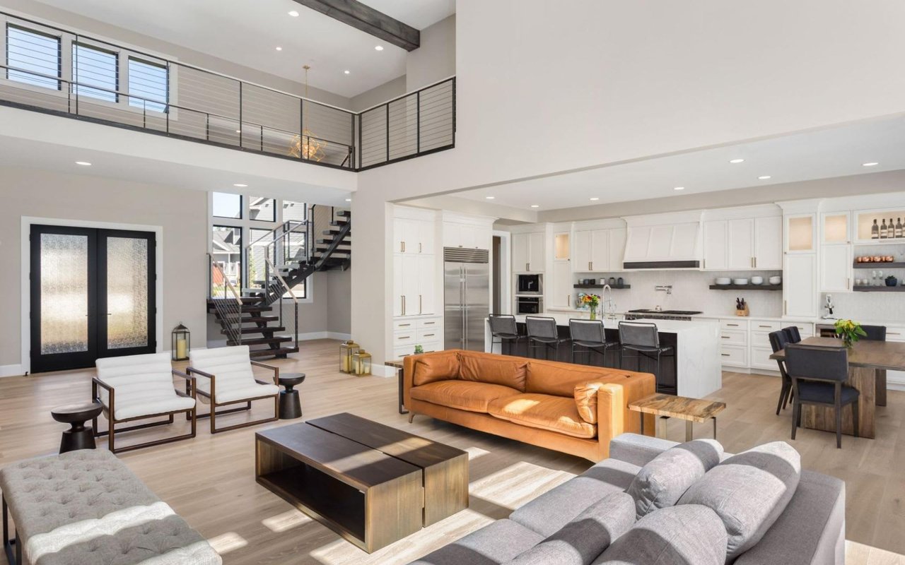 Modern open-plan home featuring a tan leather couch, grey sofa, white kitchen with an island, high ceilings, black metal stairs, and a loft walkway.