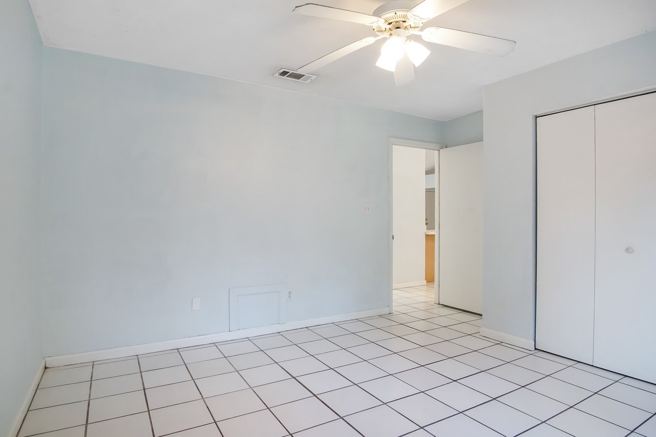 Empty room with light blue walls, white tile floor, ceiling fan, and closed closet doors. Open door reveals a glimpse into another room.