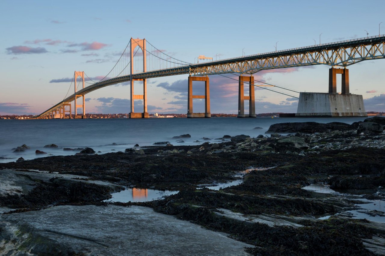 Newport's Claiborne Pell Bridge at golden hour, viewed across a rocky shoreline.