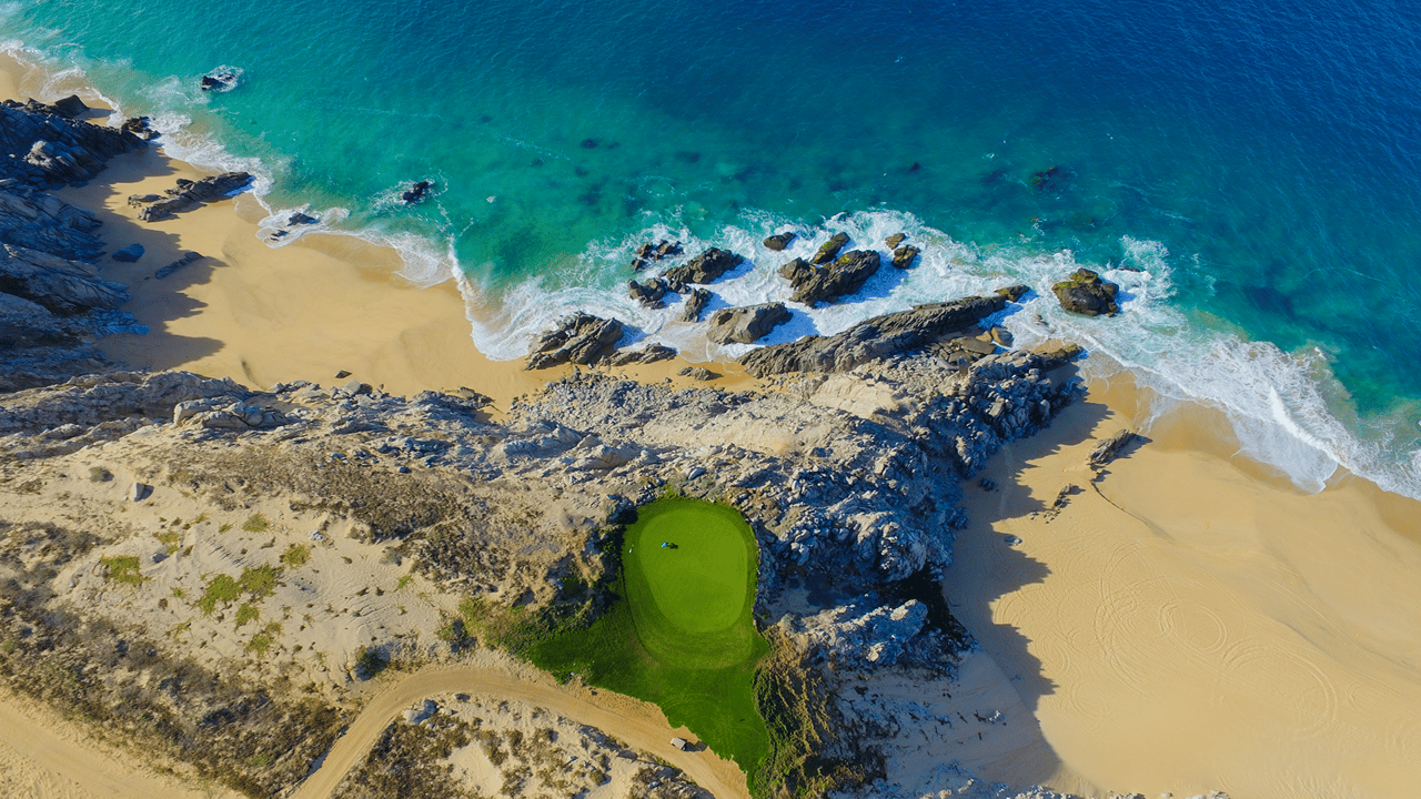 A golf green sits between rocky shoreline and turquoise surf.
