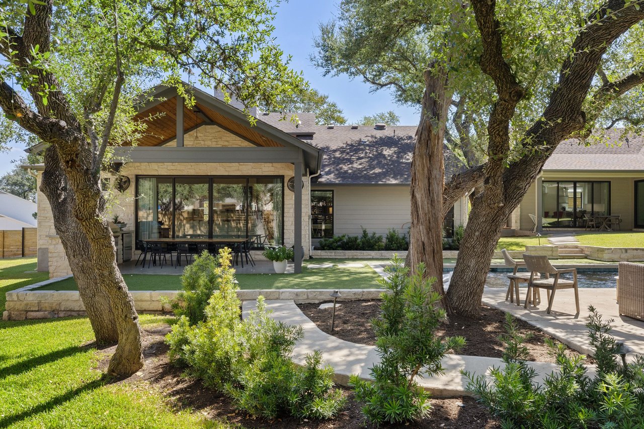 Home’s rear patio with covered outdoor dining, mature trees, landscaped lawn, and pool.