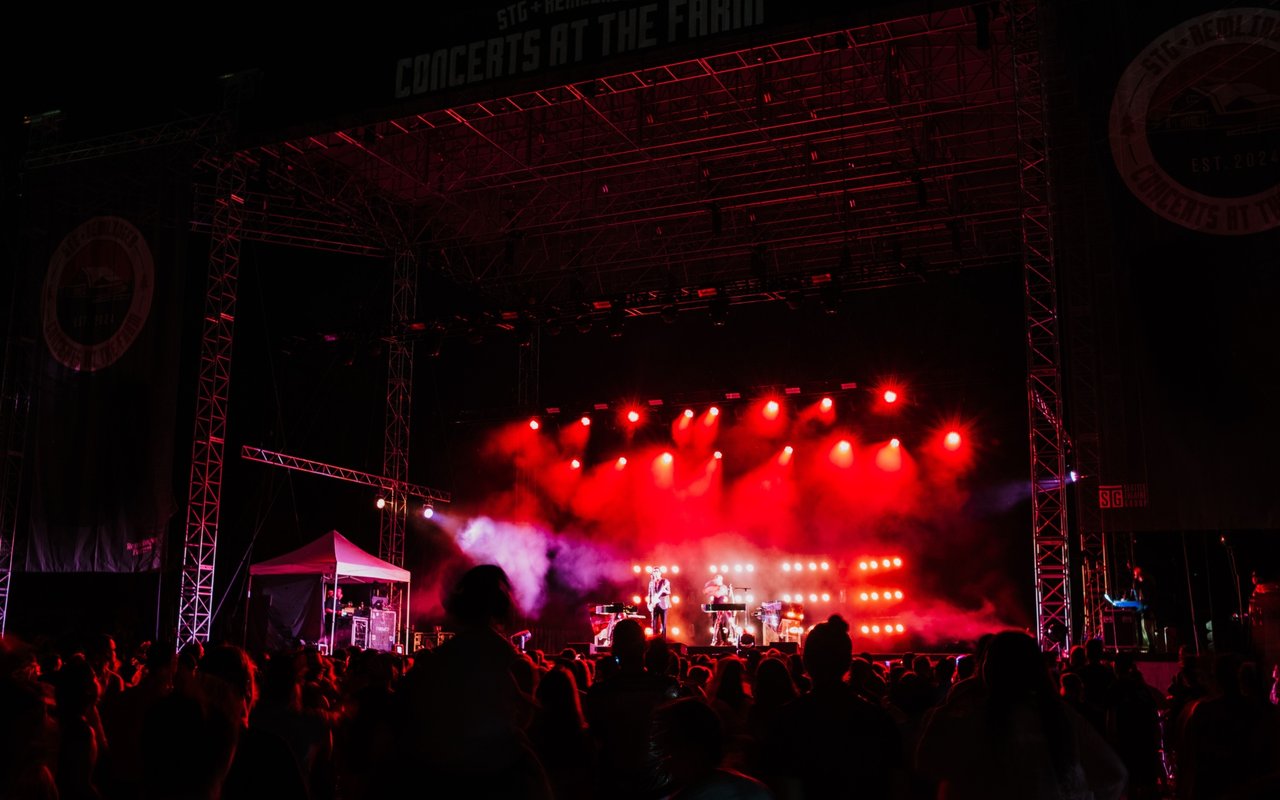 Musicians perform beneath red stage lights as a crowd watches at an outdoor concert.
