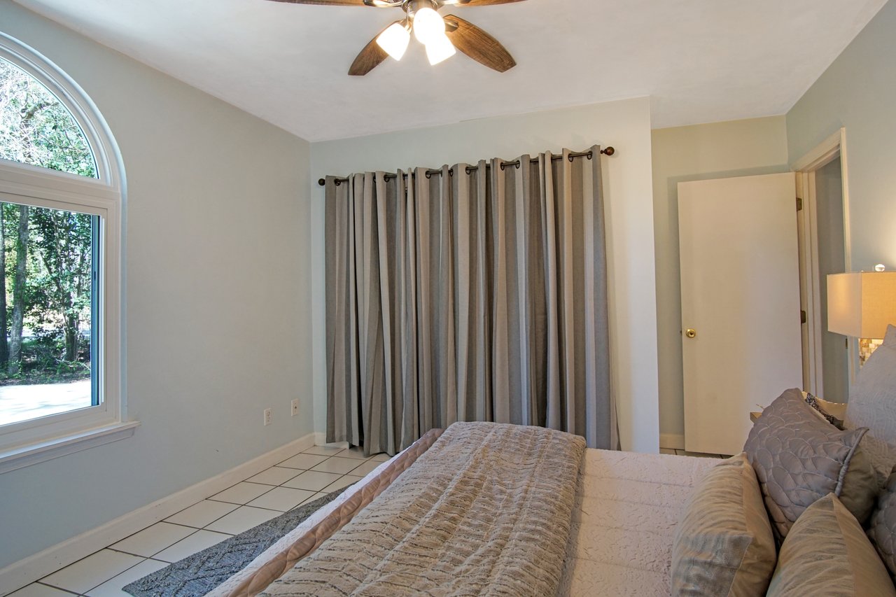 A tranquil bedroom with light blue walls, featuring a large window and arched curtains. A bed with gray bedding and soft pillows adds coziness.