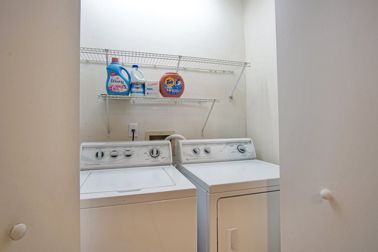 Laundry room with a white washer and dryer set below a wire shelf holding detergent, bleach, and fabric softener. Bright, clean, and organized space.