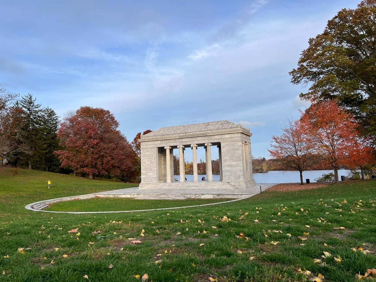 Stone pavilion beside a pond, surrounded by autumn trees in a grassy park.