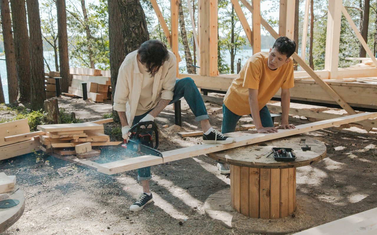 Two people cut lumber while building a wooden outdoor structure among trees.