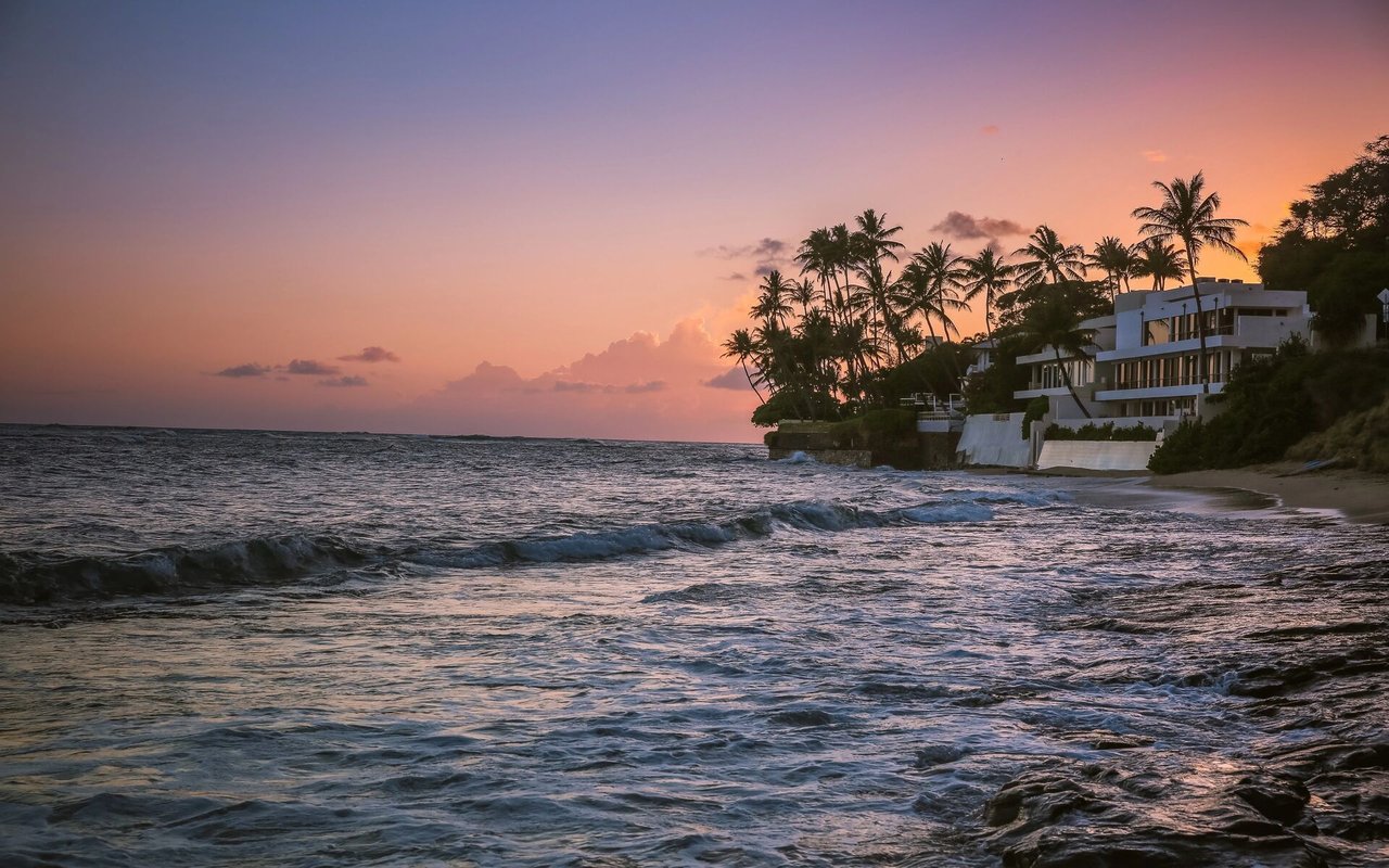 Sunset over a tropical coastline, with waves and palm-lined homes along the shore.