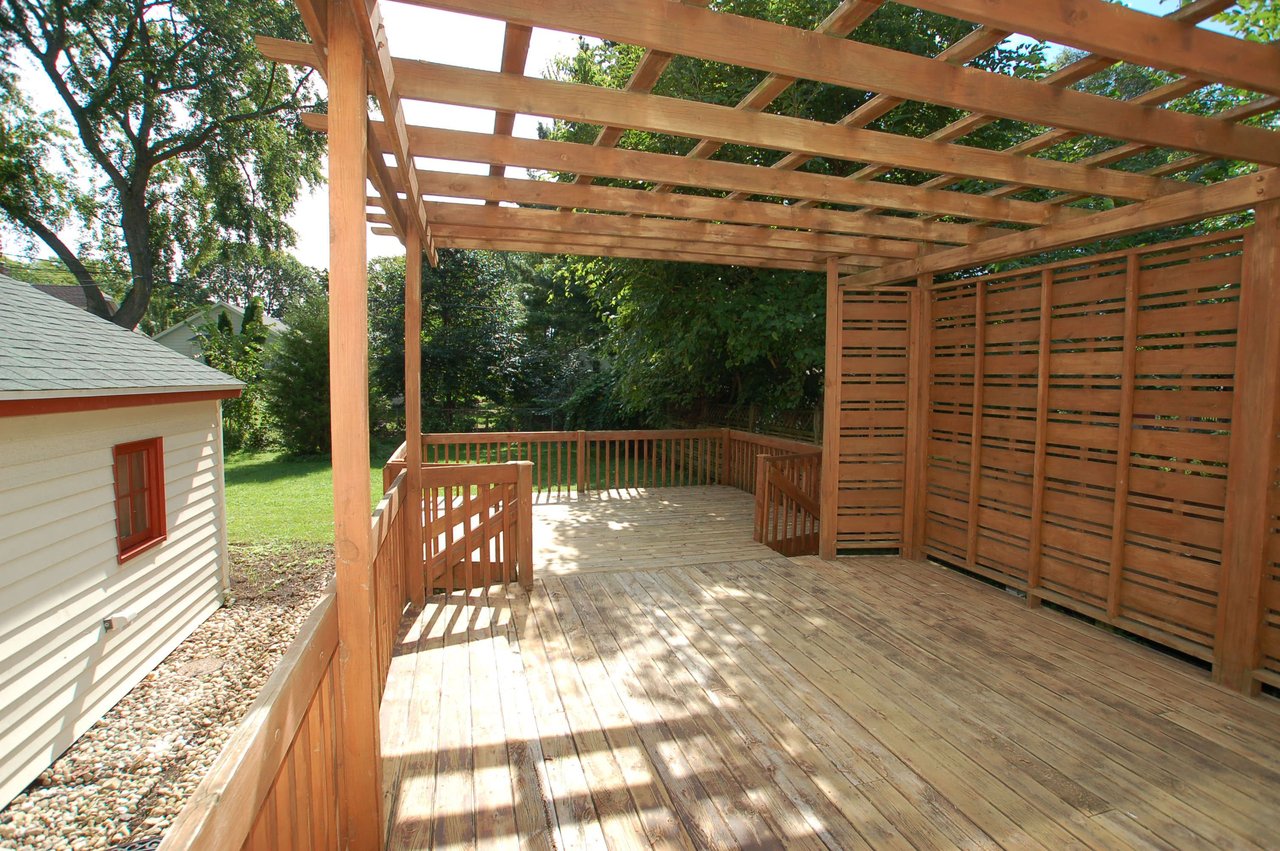 Wooden backyard deck beneath a pergola, with a slatted privacy screen and leafy trees.