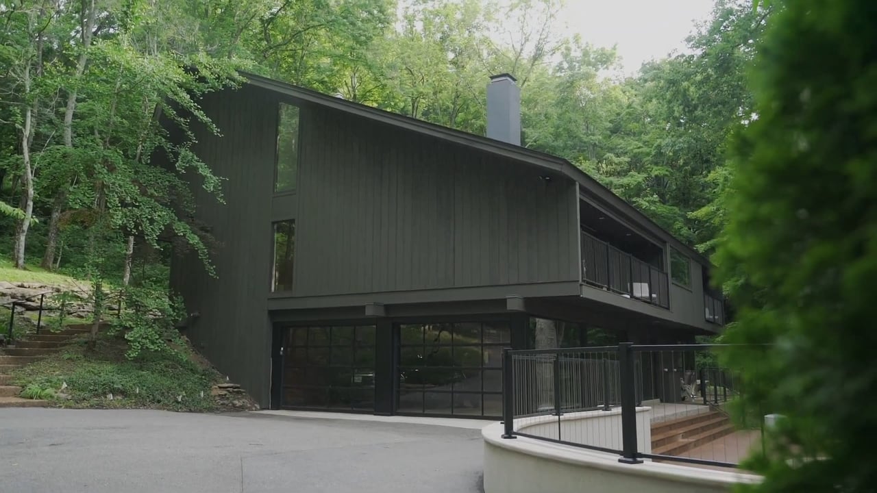 Modern dark-sided home among trees, with a sloped roof, large glass doors, and raised deck.