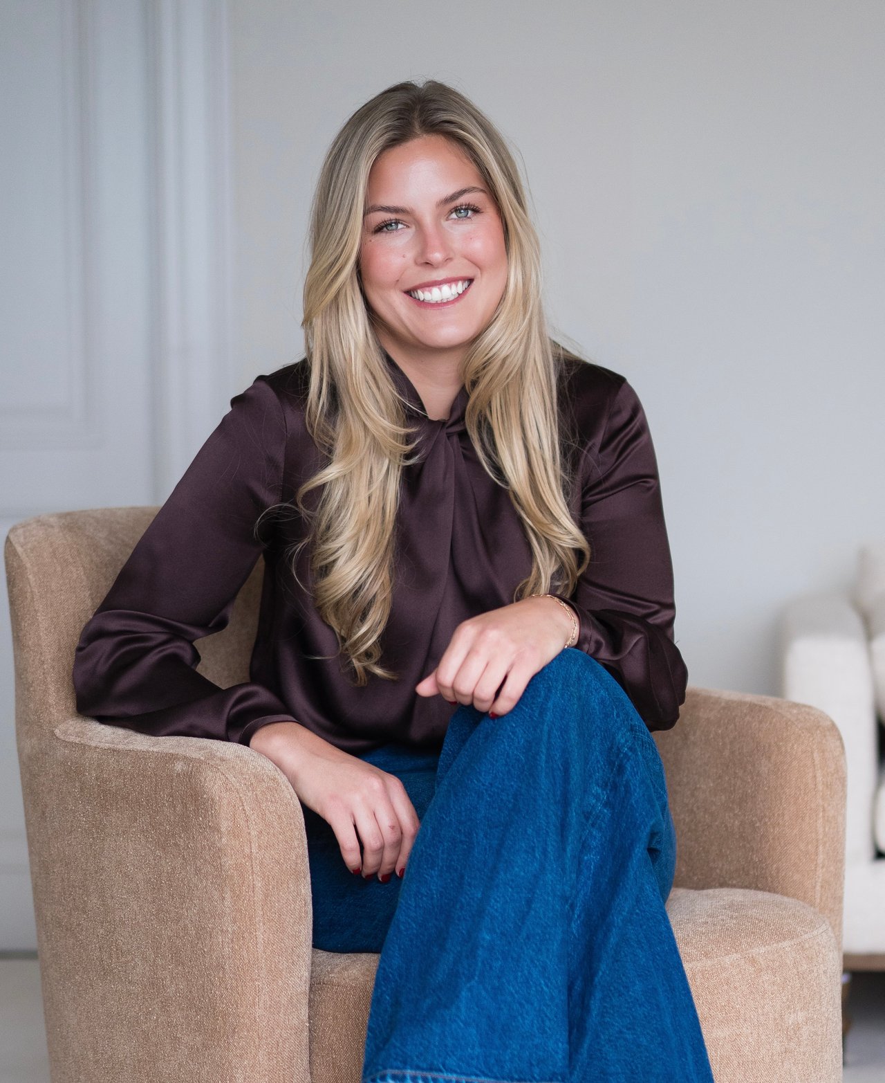 Tatum Shaver seated in a beige armchair, smiling in a dark blouse and blue pants.