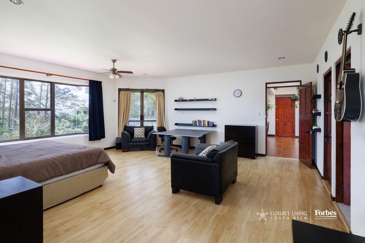 Furnished bedroom with large windows, a work table, dark sofas, and guitars mounted on the wall.