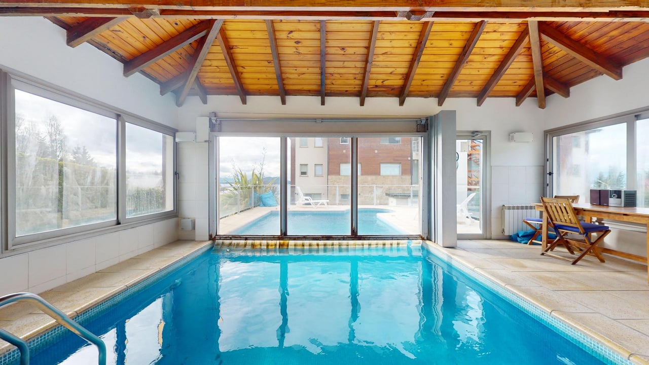 Indoor swimming pool beneath a vaulted wooden ceiling, with sliding doors opening to an outdoor pool.