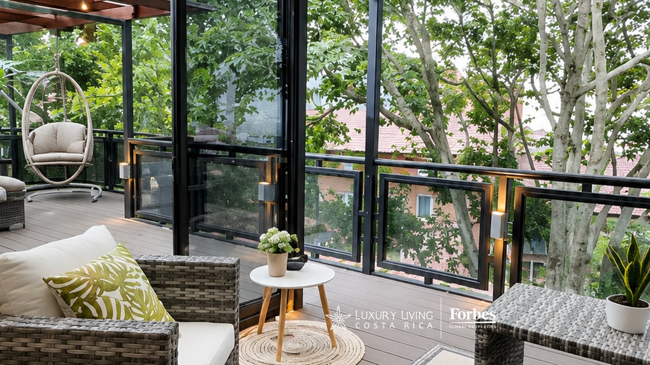 Covered balcony with wicker seating and glass railings overlooking leafy trees.