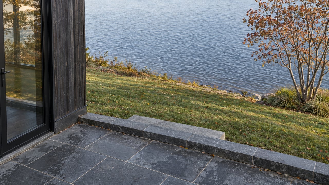 Stone terrace overlooks sloping lawn and lake beside dark cedar siding and a sparse-leaved tree.