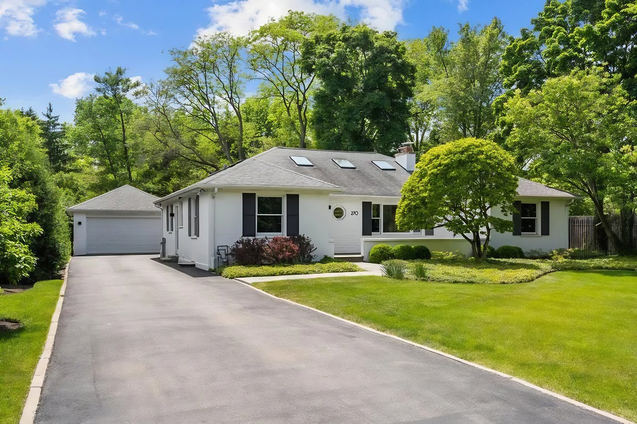 White single-story home with roof skylights, dark shutters, and a broad driveway framed by mature trees.