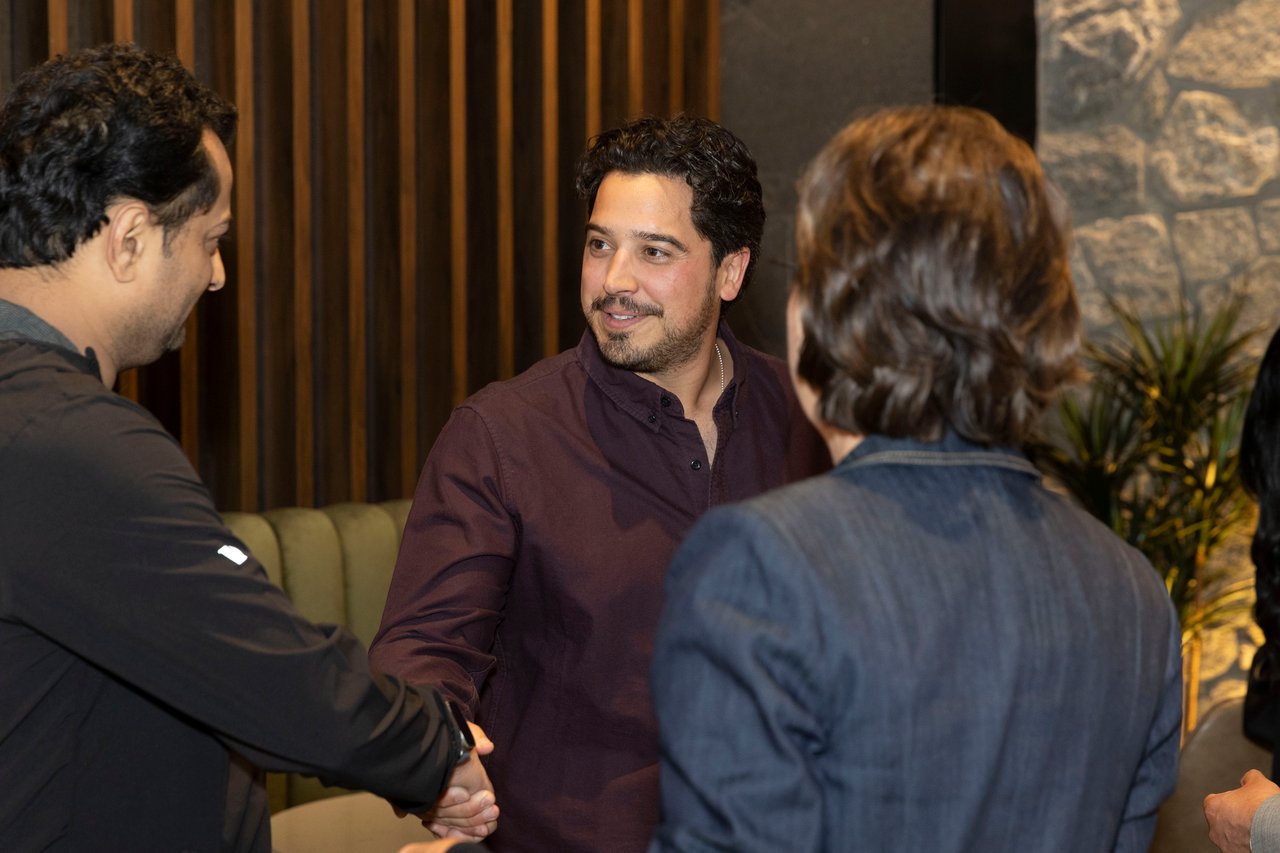 Guests greet one another at an indoor event; a man in burgundy shakes hands with another guest.