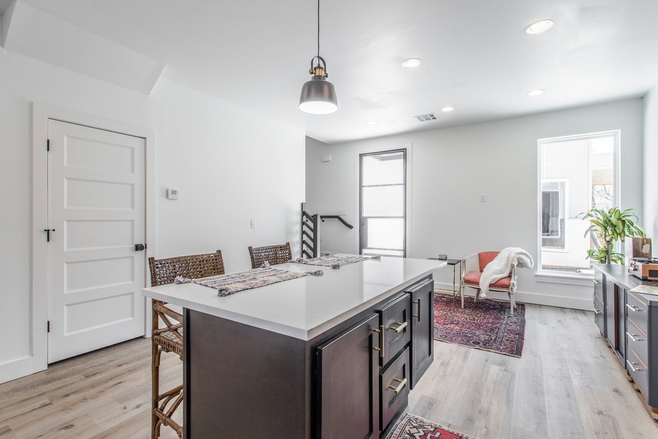 Kitchen with a large dark island, woven chairs, and a sitting area by tall windows.