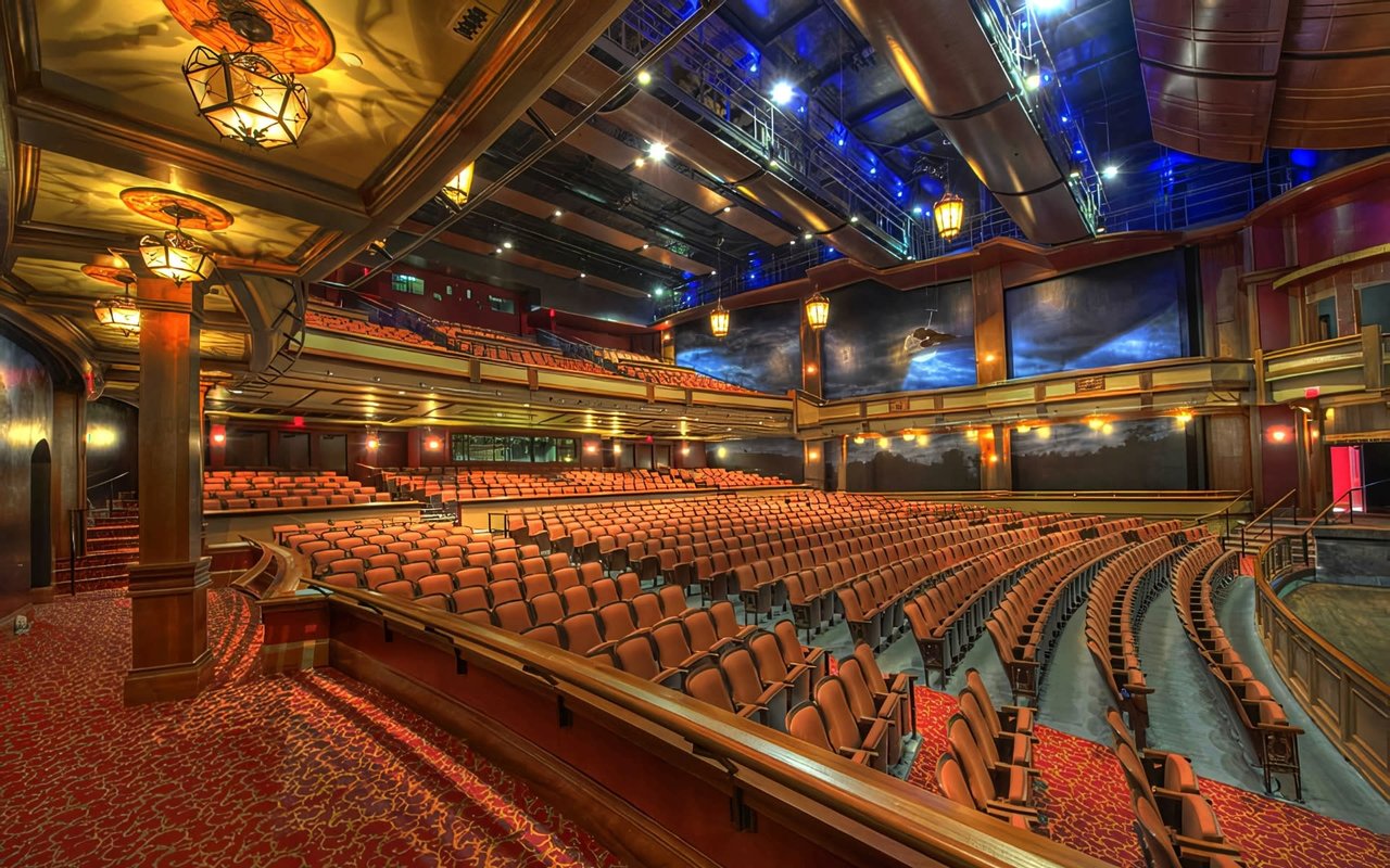 Empty grand theater featuring rows of orange seats, patterned red carpet, wooden columns, hanging lanterns, and overhead stage rigging with blue lights.