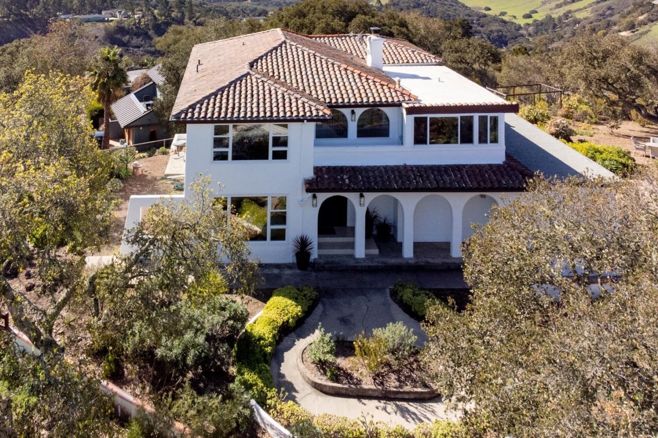 White two-story home with a tiled roof, arched porch, and curved driveway amid mature trees.