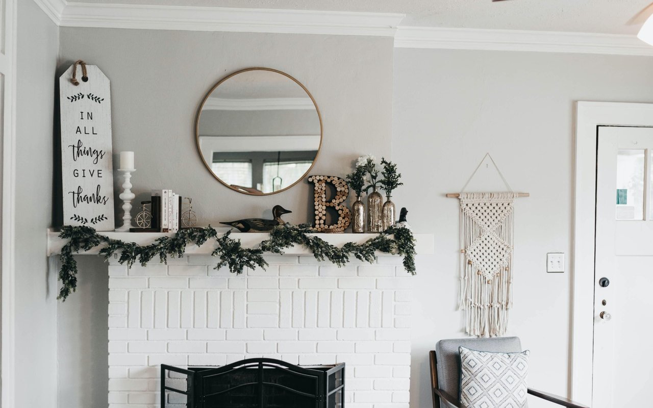 Living room with a white brick fireplace, evergreen mantel garland, round mirror, and gray chair.